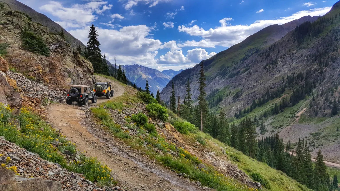 Two off-road vehicles on a mountain trail with a scenic view of mountains and blue sky.